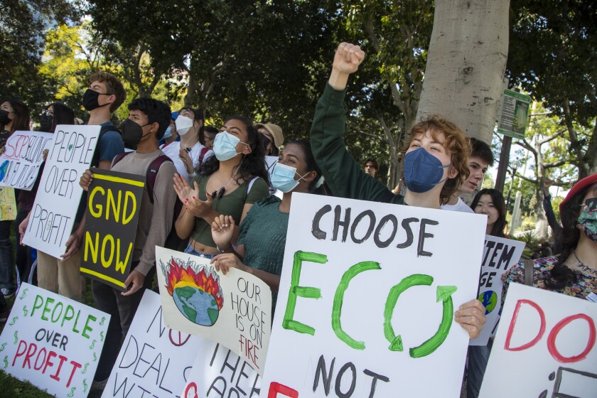 A group of young people in masks stand together beneath trees, clapping and holding posters.