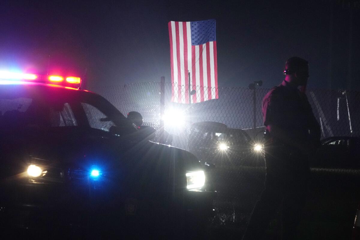 A man stands next to a police vehicle with lights on, near an illuminated U.S. flag