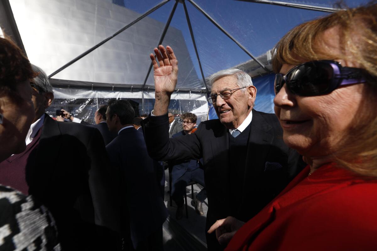 Eli Broad waves among a crowd under a transparent tarp