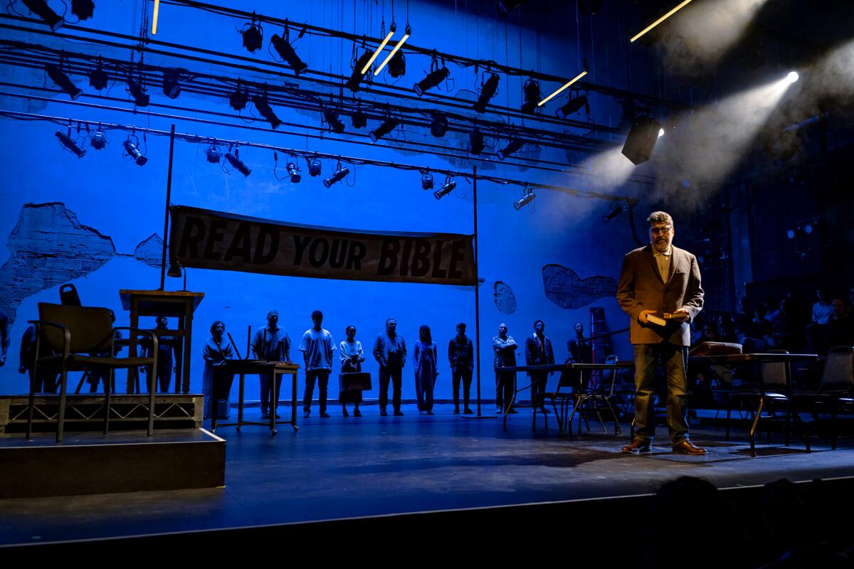 An actor is illuminated at the front of a stage, with other actors in blue light lined up behind him.