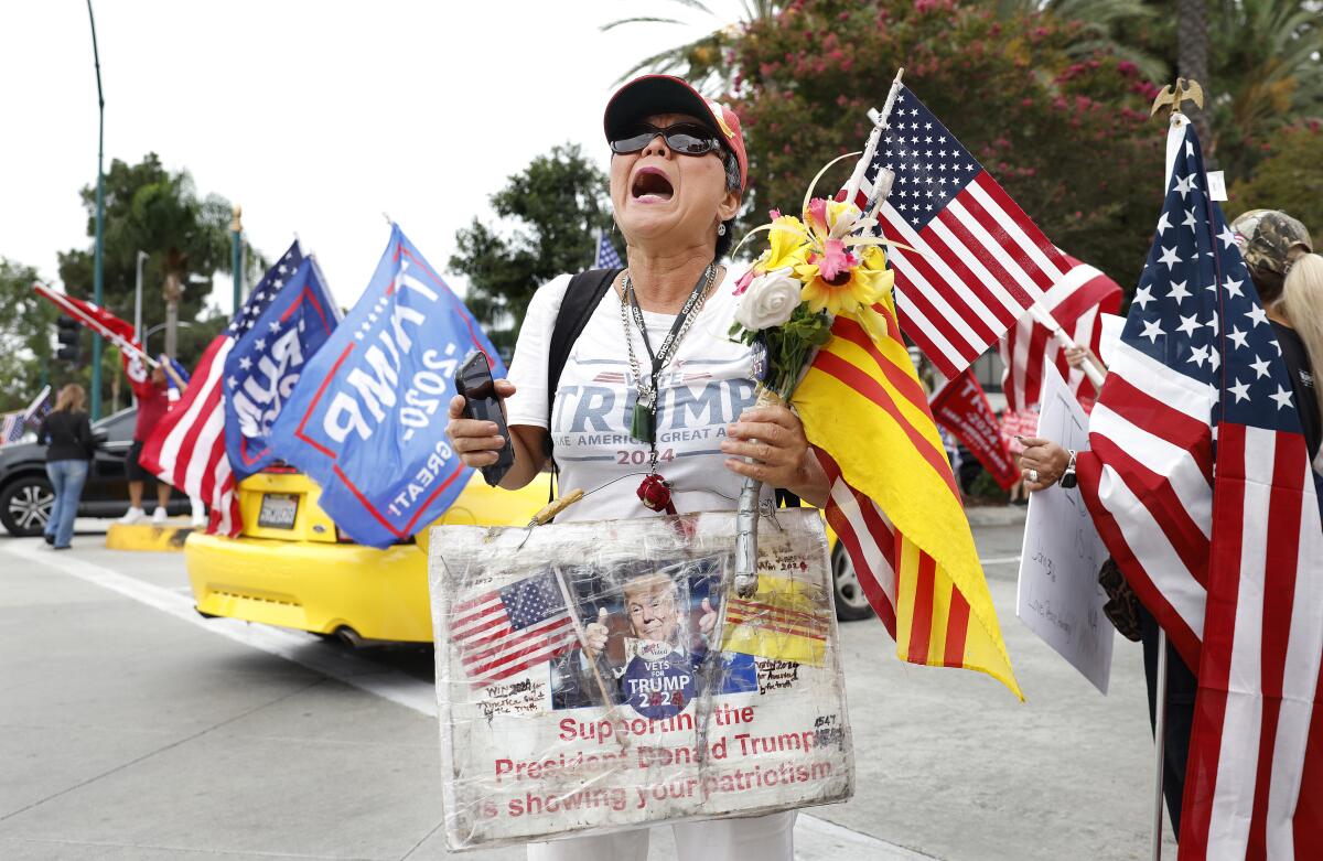 A person with a white shirt bearing the word Trump holds flowers and flags near more U.S. flags and a blue Trump banner 