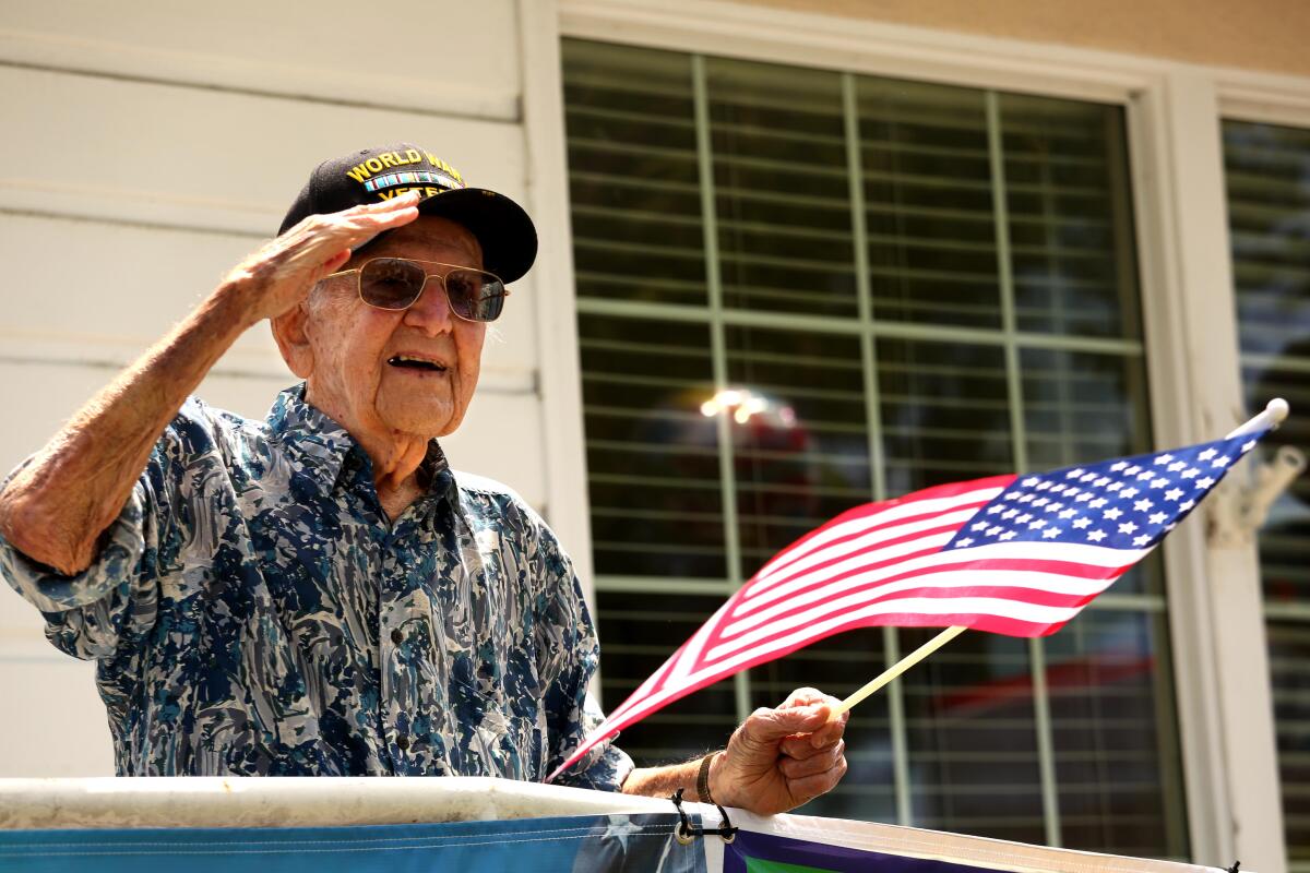 An elderly man wearing a baseball cap salutes while holding an American flag