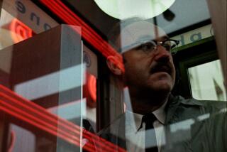 A paranoid man looks out of a glass phone booth.