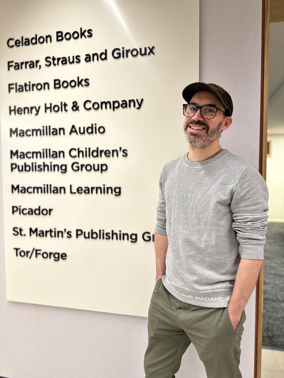 Macmillan Publishing'in izlerini listeleyen bir panonun önünde duran şapkalı ve uzun süveterli bir adam.