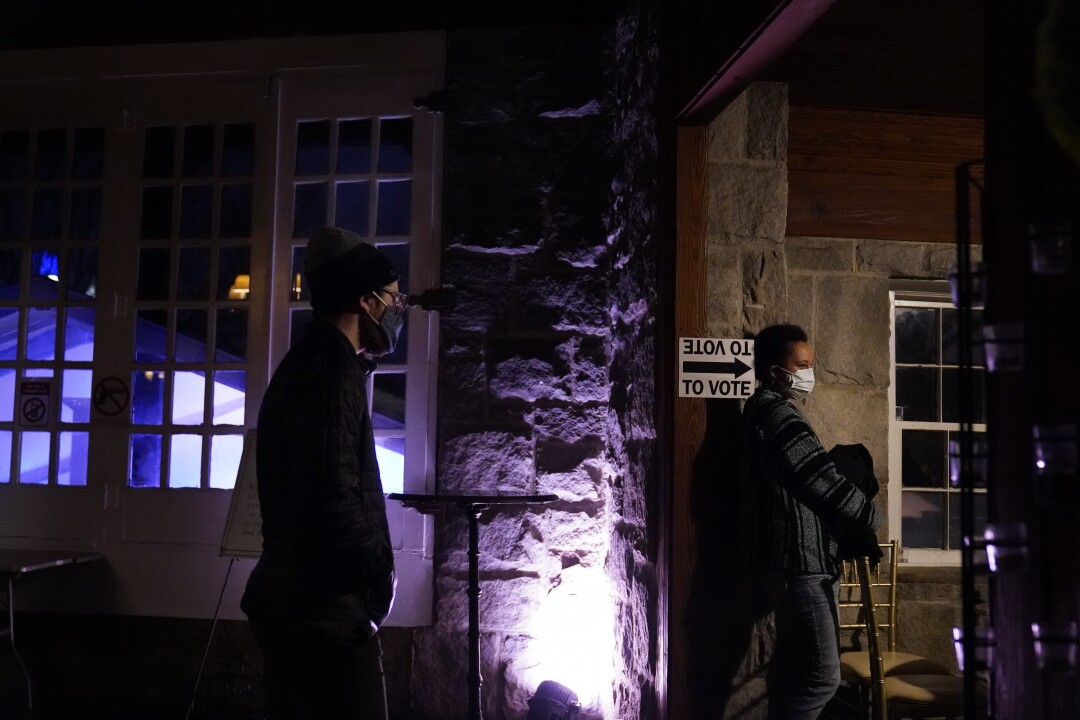 Next to a "To vote" sign with an arrow, two voters stand in line, a few feet of space between them, after dark.
