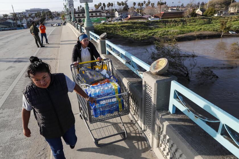 Pajaro, CA, Thursday, March 16, 2023 - Maria Terriquez, right, and Blanca Garcia push a shopping cart of supplies across the Pajaro River Bridge. They are two of dozens of residents who defied evacuation orders and remain in their homes nearly a week after a levee break flooded their community. (Robert Gauthier/Los Angeles Times)