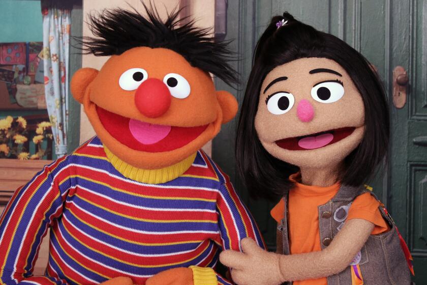 An orange muppet with tousled black hair and a striped shirt smiles with a muppet resembling a girl with dark brown hair.