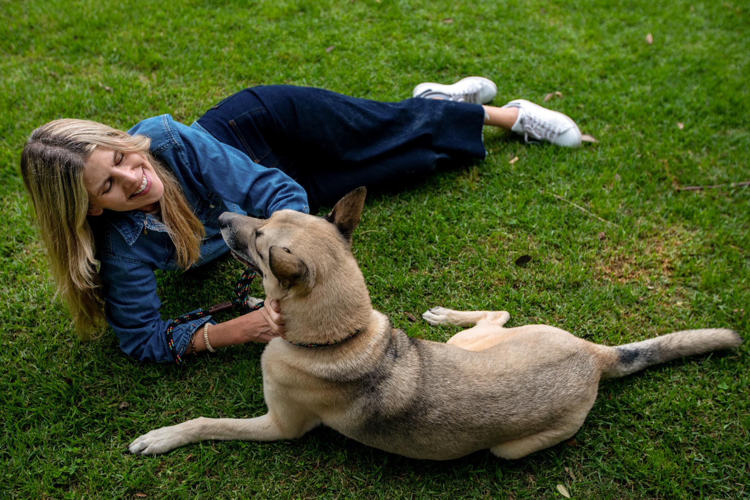 A woman lies on the grass with her dog. They are looking at each other.