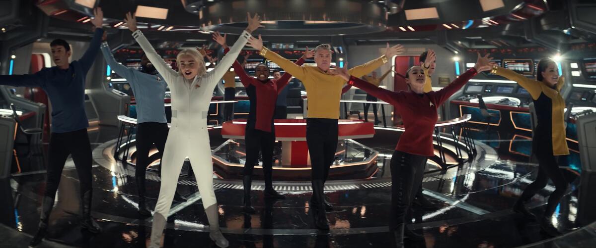 Seven people stand with their arms and legs spread outward on the command deck of a spaceship.