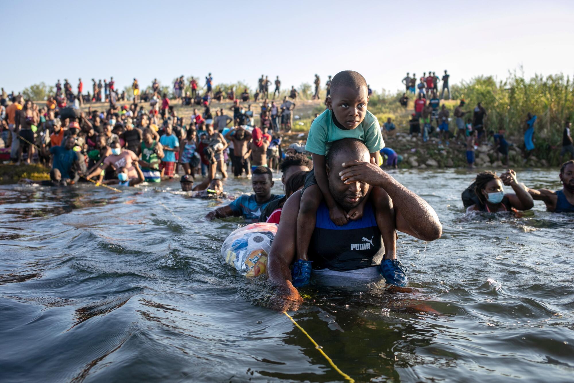 A man shields his face as he crosses the Rio Grande with a youngster on his shoulders.