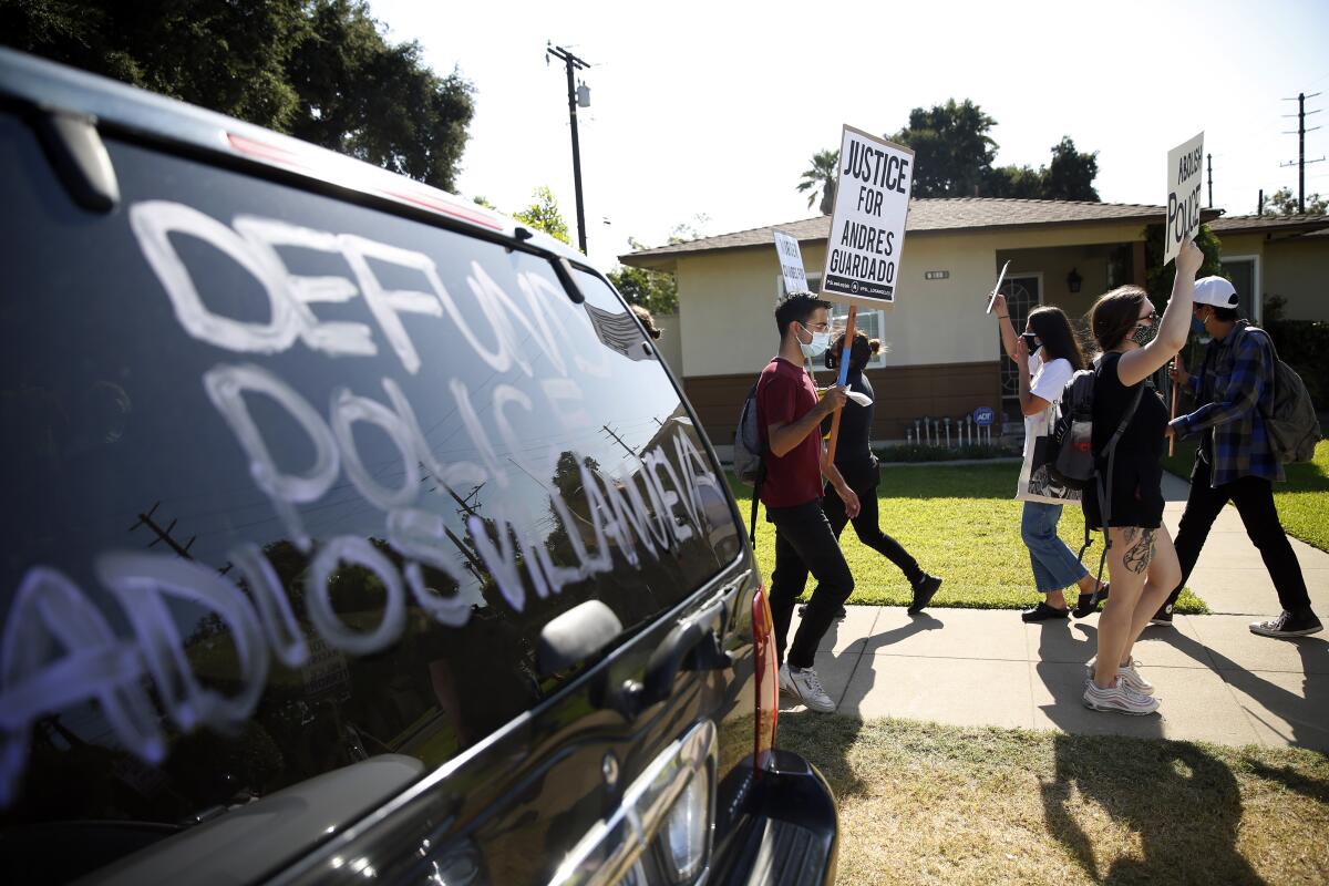 Masked protesters holding signs on a sidewalk beside a vehicle with "DEFUND POLICE" on its window