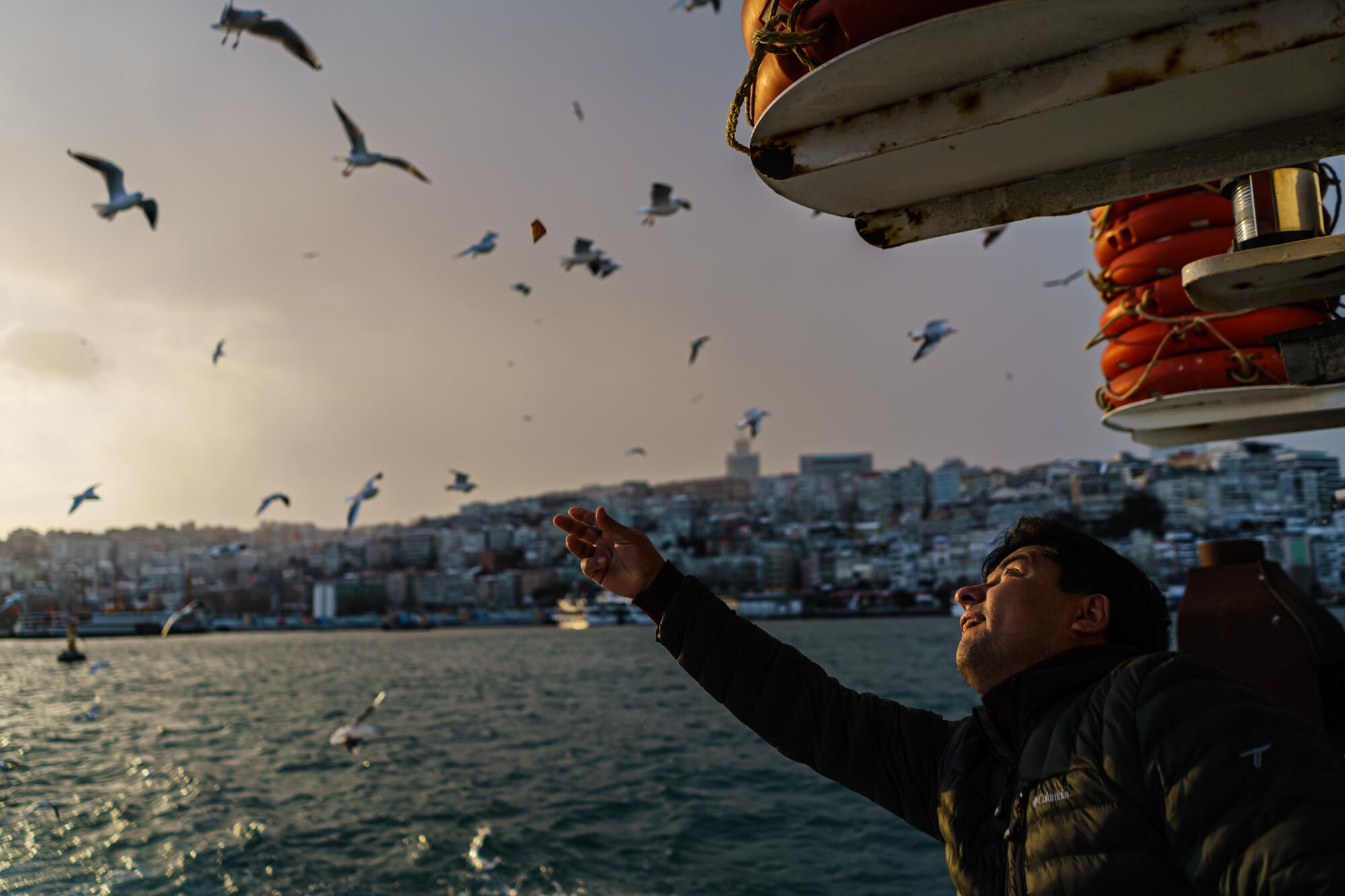 A man on a boat extends his hand against a backdrop of buildings and a flock of birds over the water