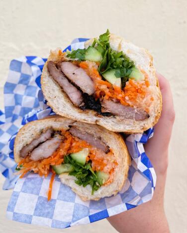 A vertical photo of a hand holding two stacked halves of a roast pork banh mi from Bé ? restaurant in Silver Lake.