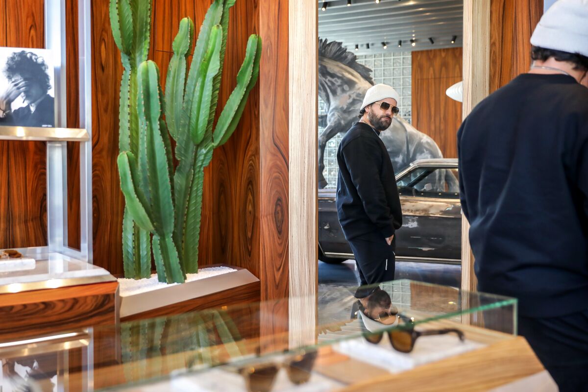 A man trying on sunglasses looks in a mirror in a store with wooden shelves and a cactus. 