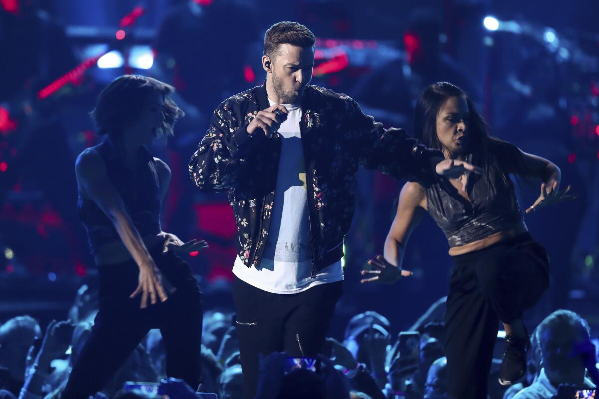 Justin Timberlake in a white shirt and camouflage jacket holding a microphone and dancing on a dark stage with two women