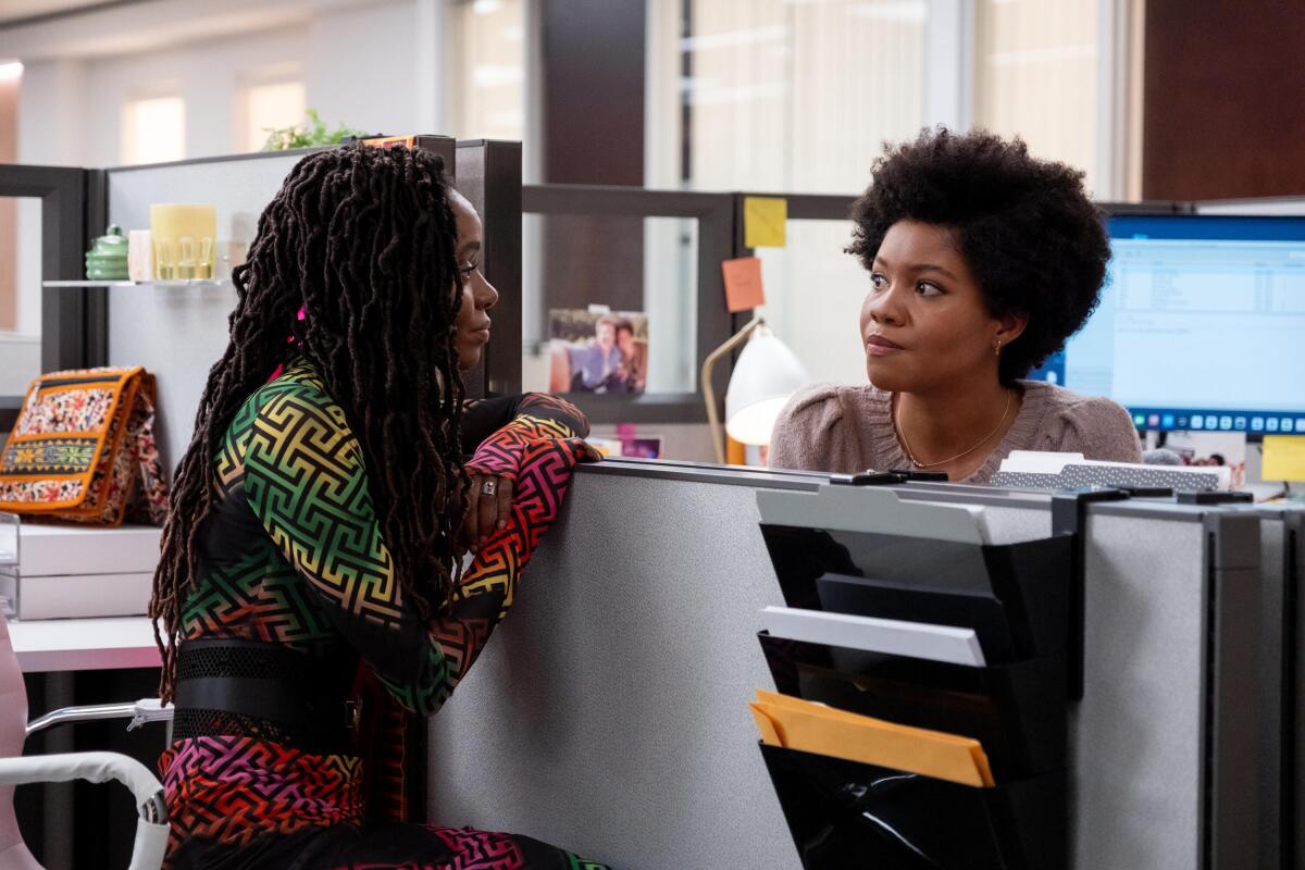 Two Black women talking over an office cubicle divider.
