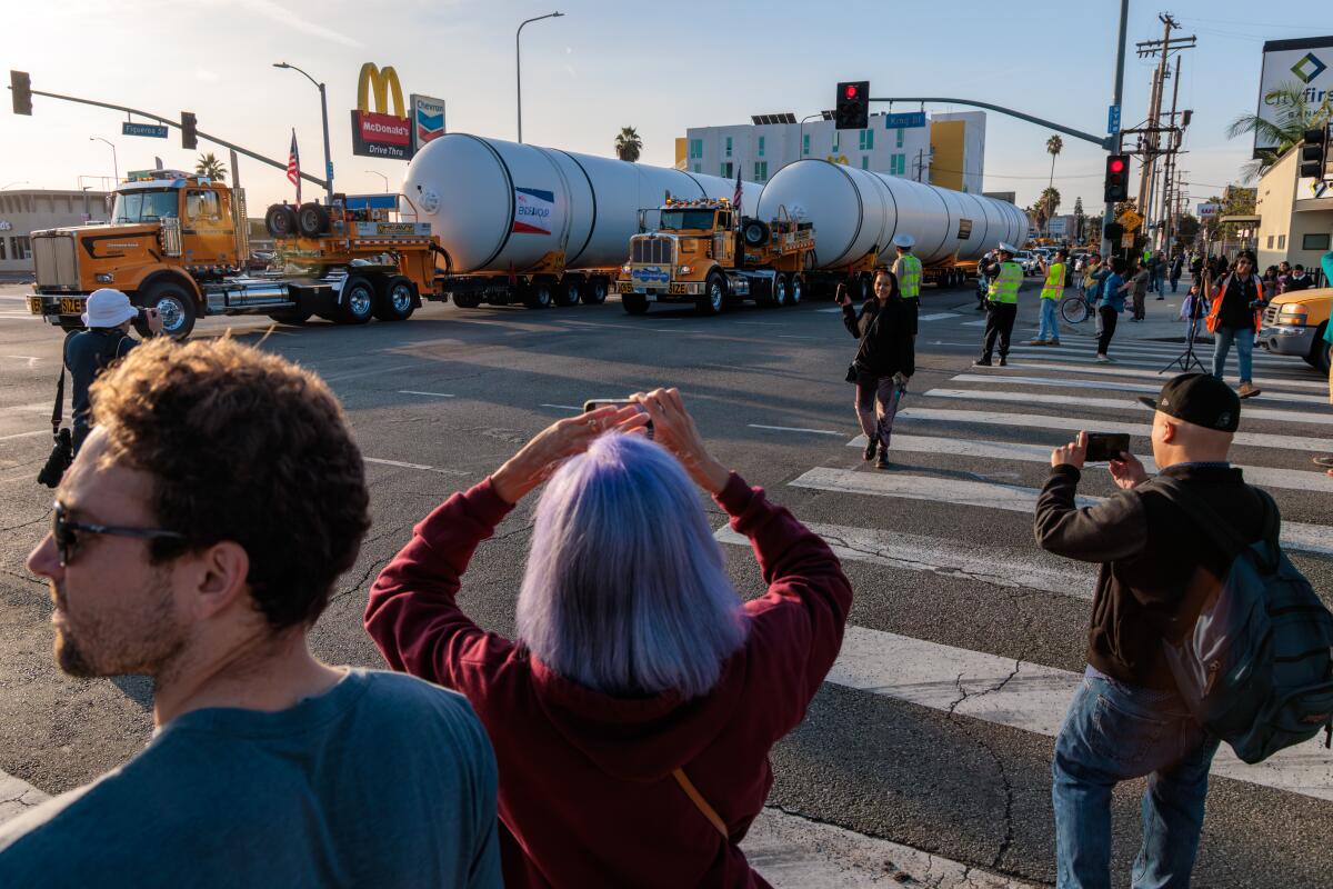 People take pictures as two long metal cylinders are trucked along a street.