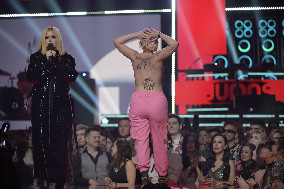 A blond woman in a long black coat at a microphone onstage as a topless woman in pink pants displays words on her back