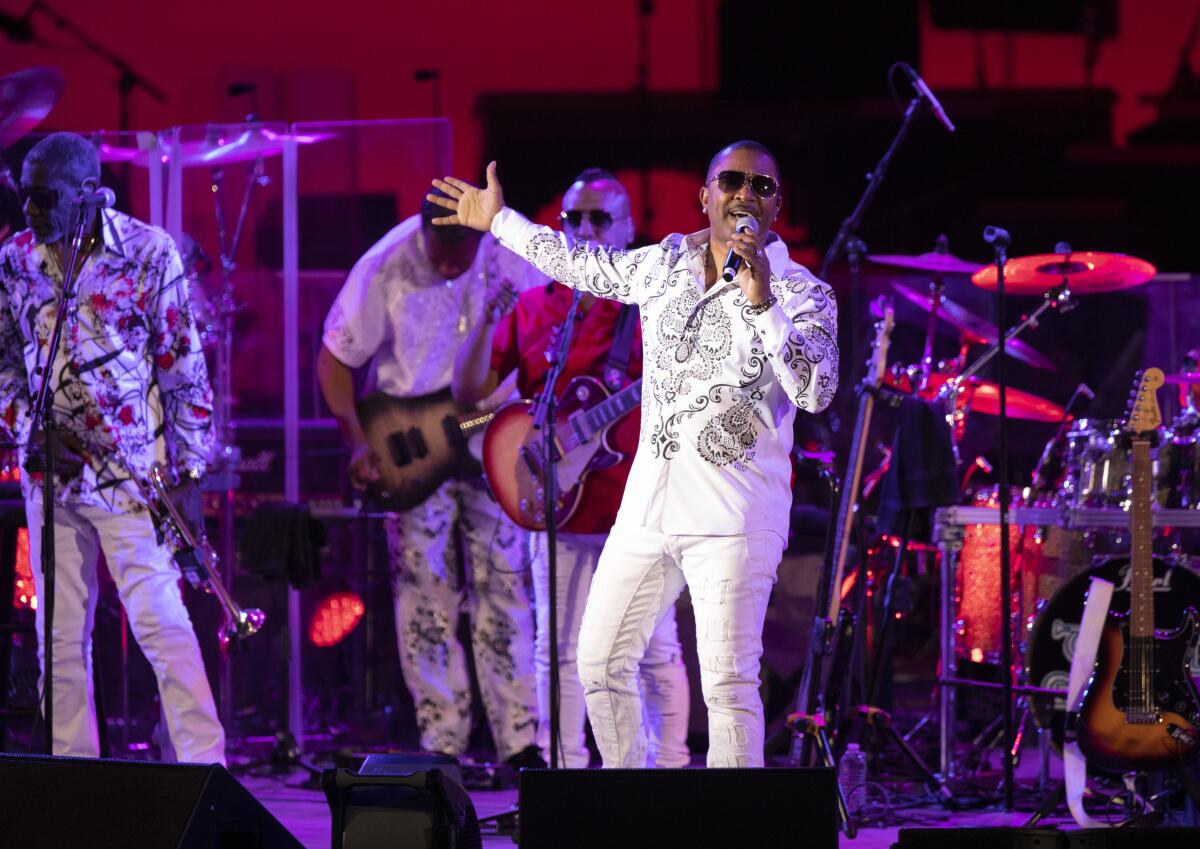 A man in sunglasses and white shirt and pants sings into a microphone on stage.