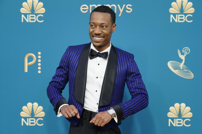 A man in a blue and black striped tuxedo jacked and black smiles with his elbows out to his sides
