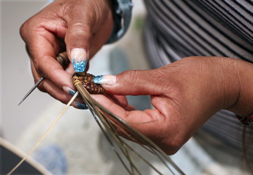 a close up of a woman's hands with sparkling blue nail polish weaving pine needles into a basket