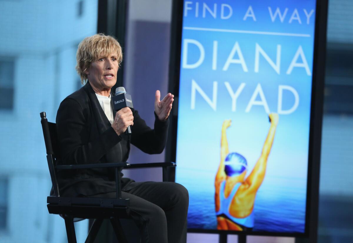 A woman speaks into a microphone next to a large image of a swimmer raising her arms and the words Find a Way Diana Nyad.