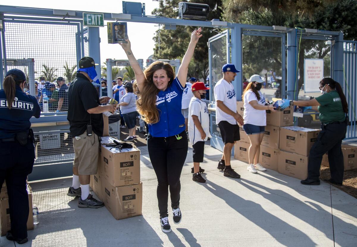 A woman in a Dodgers jersey raises both arms. In the background people go through a security check and show tickets.