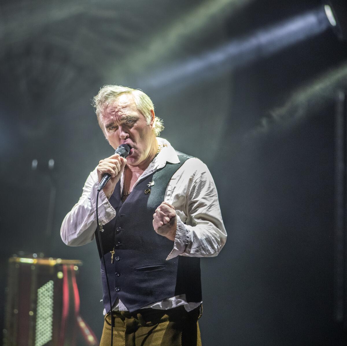 A man with white hair, wearing a vest, sings onstage