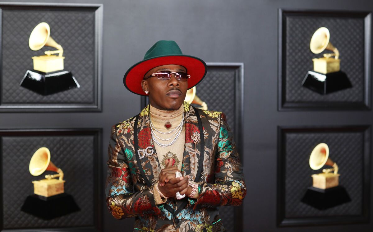 A man posing with his hands clasped in a green hat and patterned suit, surrounded by pictures of Grammy Awards.
