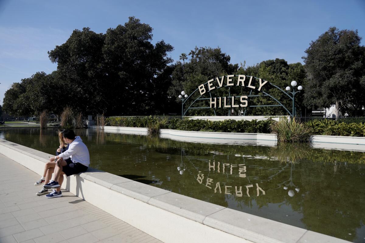 Two people sit by a pool with a "Beverly Hills" sign on the other side.