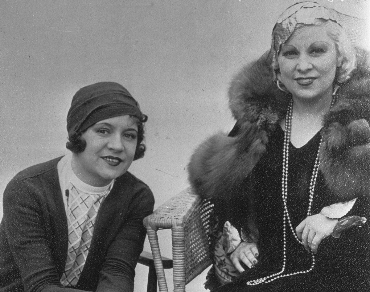 Two women sit side by side. One wears a fur and long necklaces.