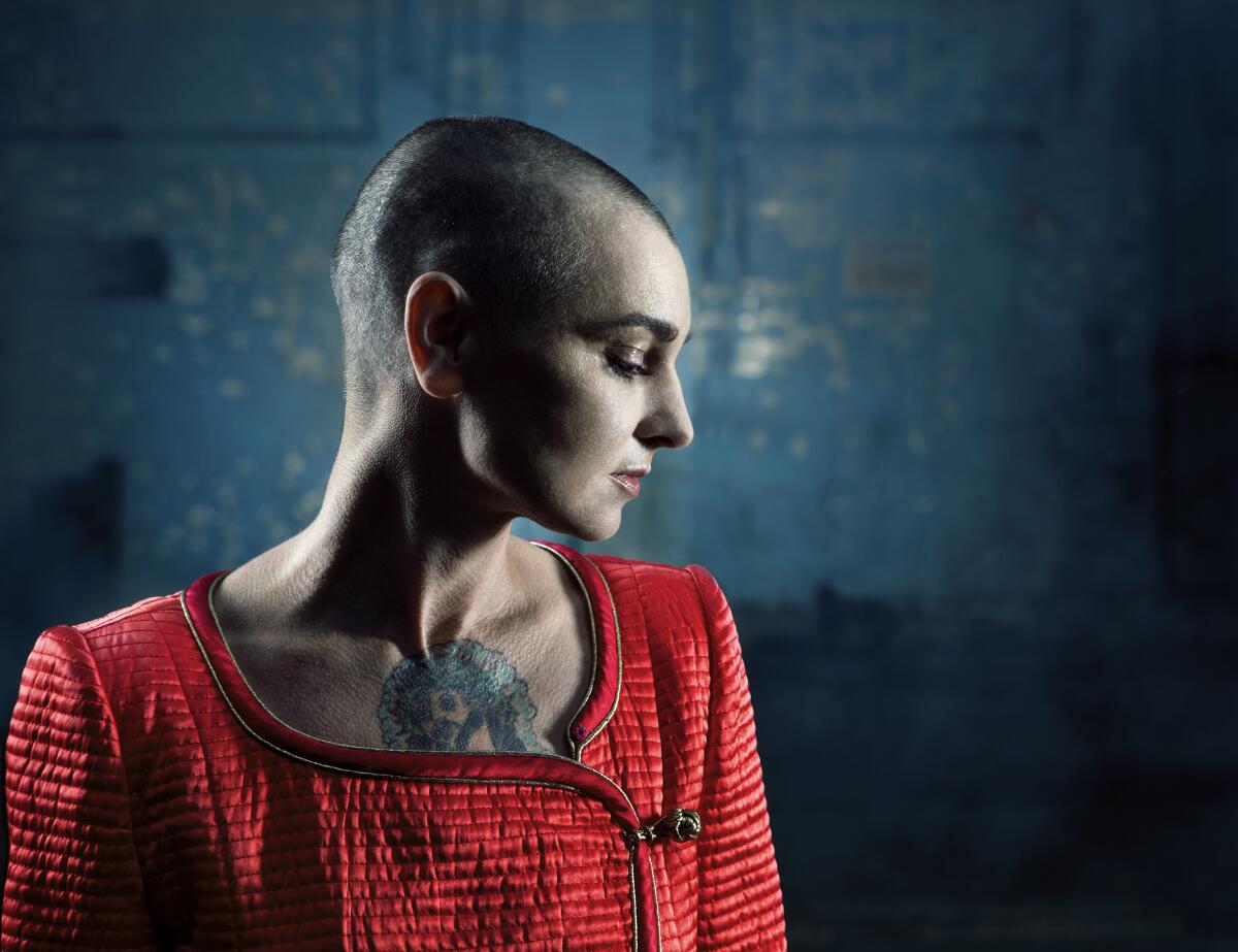 A portrait photo of a younger Sinéad O'Connor with her head shaved and a tattoo visible on her upper chest.