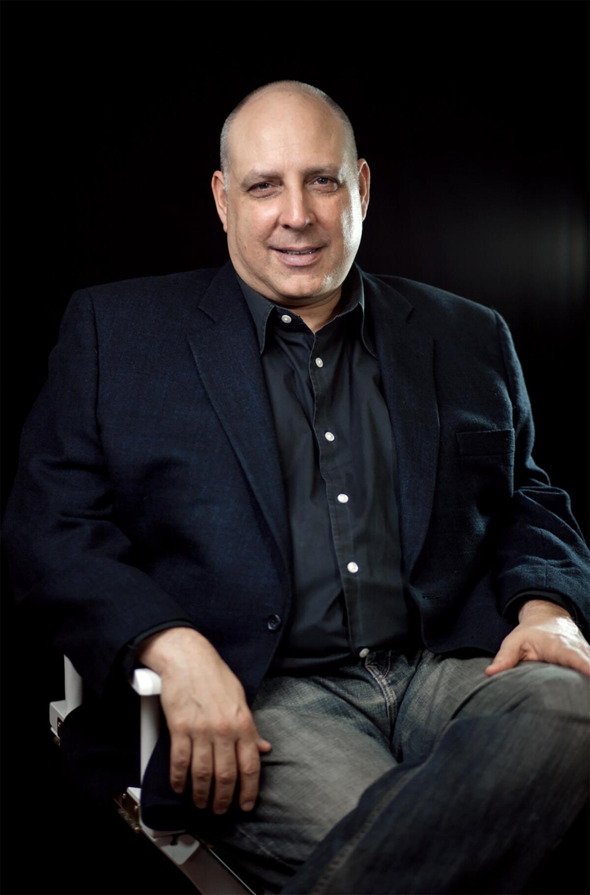 A seated, smiling bald man in a dark shirt and jacket.