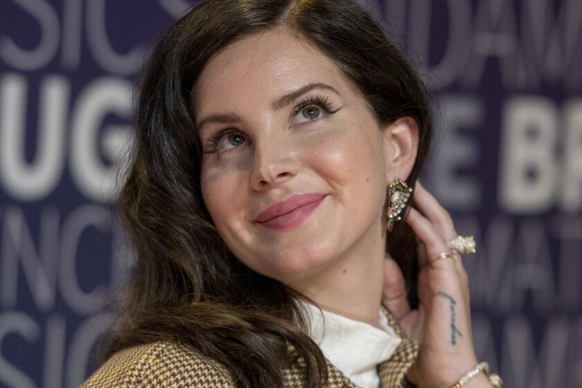 Lana Del Rey is smiling and looking up while tucking her hair behind her ear while wearing a brown plaid blazer
