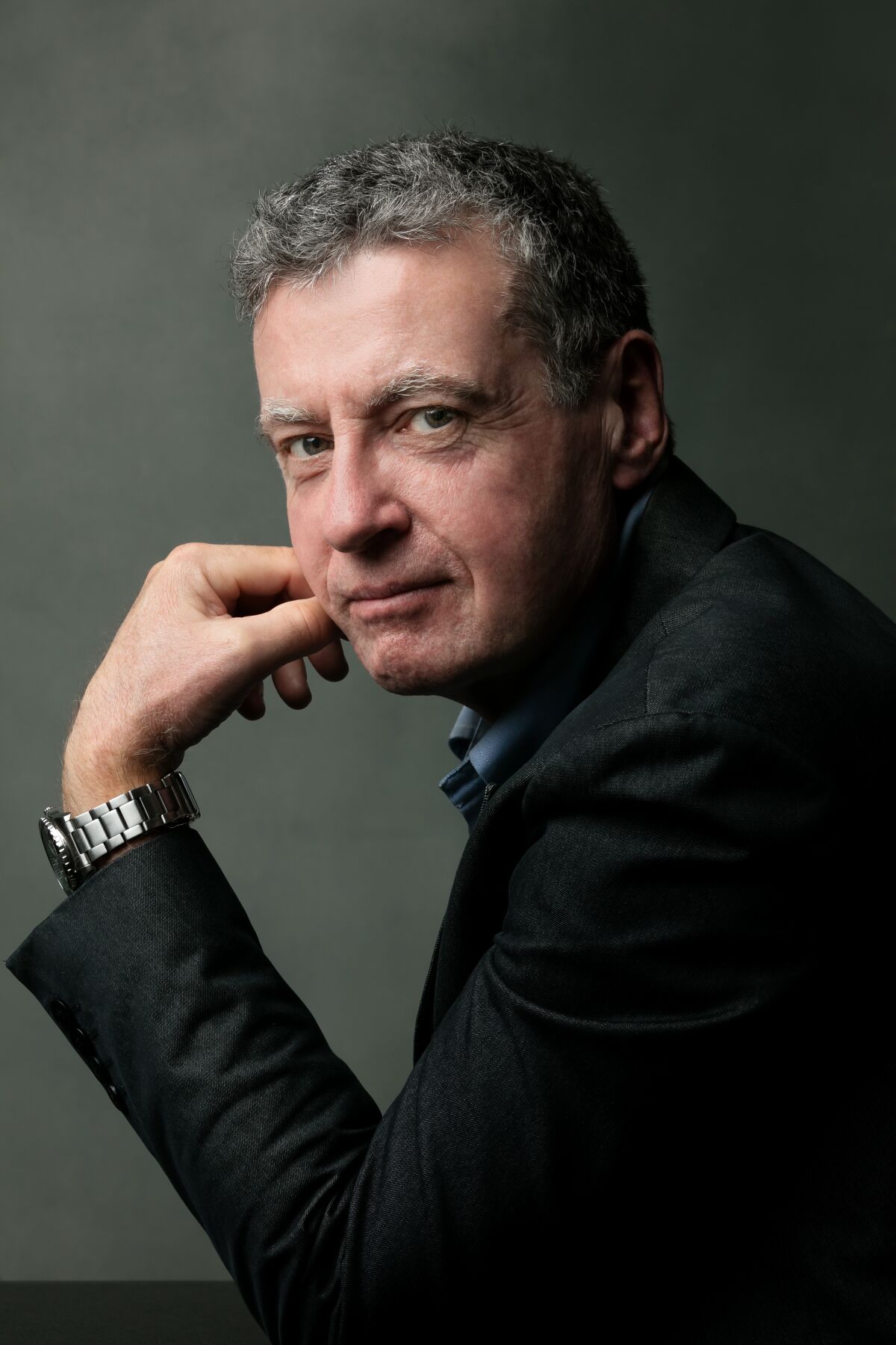 A man in a dark jacket sits with his left hand against his right cheek, looking toward the camera.