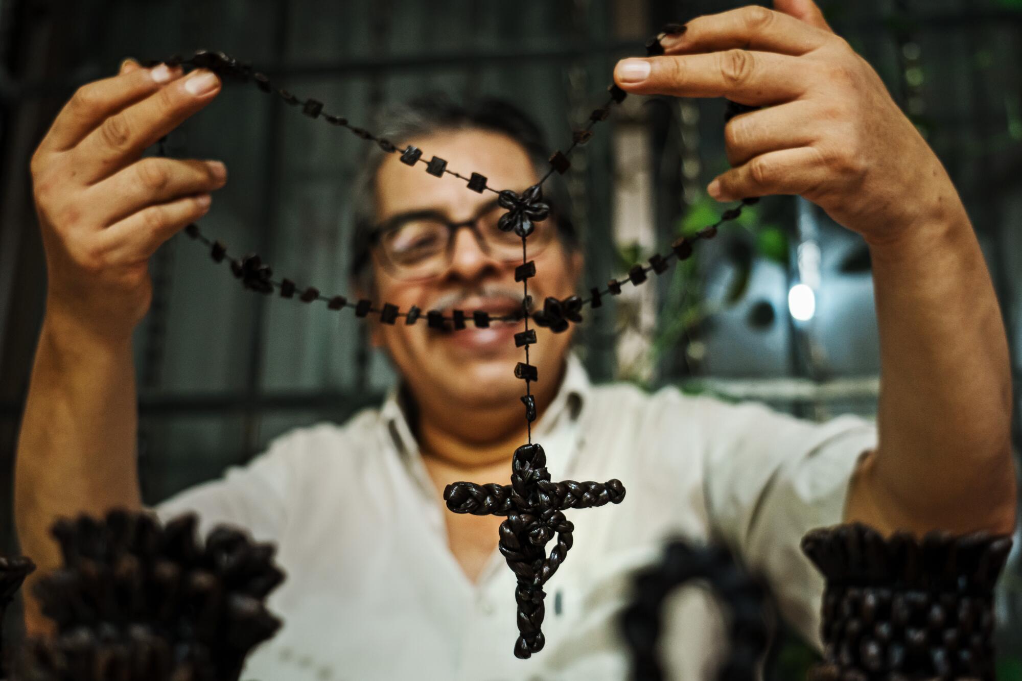 A smiling man wearing glasses holds up a necklace with a cross made out of vanilla beans 