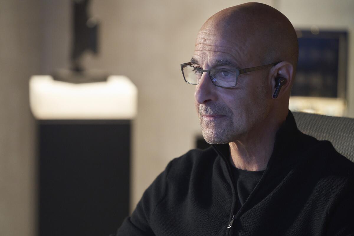 Stanley Tucci wears glasses and earbuds in a minimalist room.