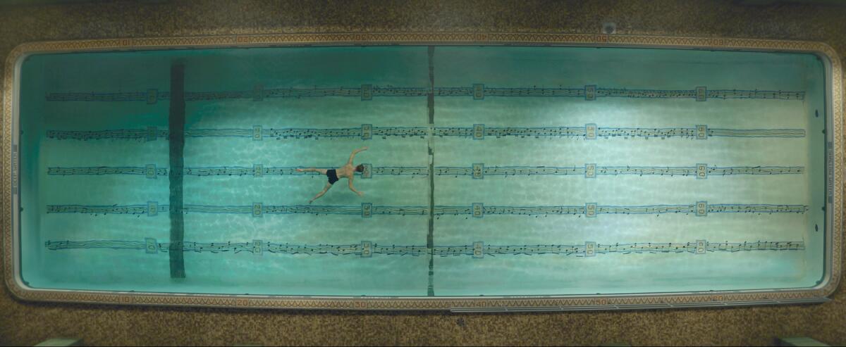 An aerial shot of a man in a pool with sheet music as lane lines