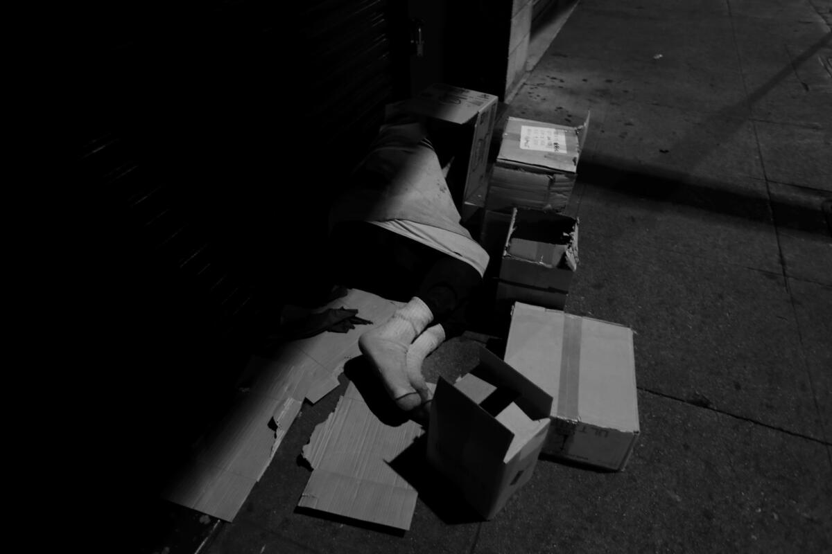 A person sleeps on a piece of cardboard, surrounded by boxes on the sidewalk on skid row.