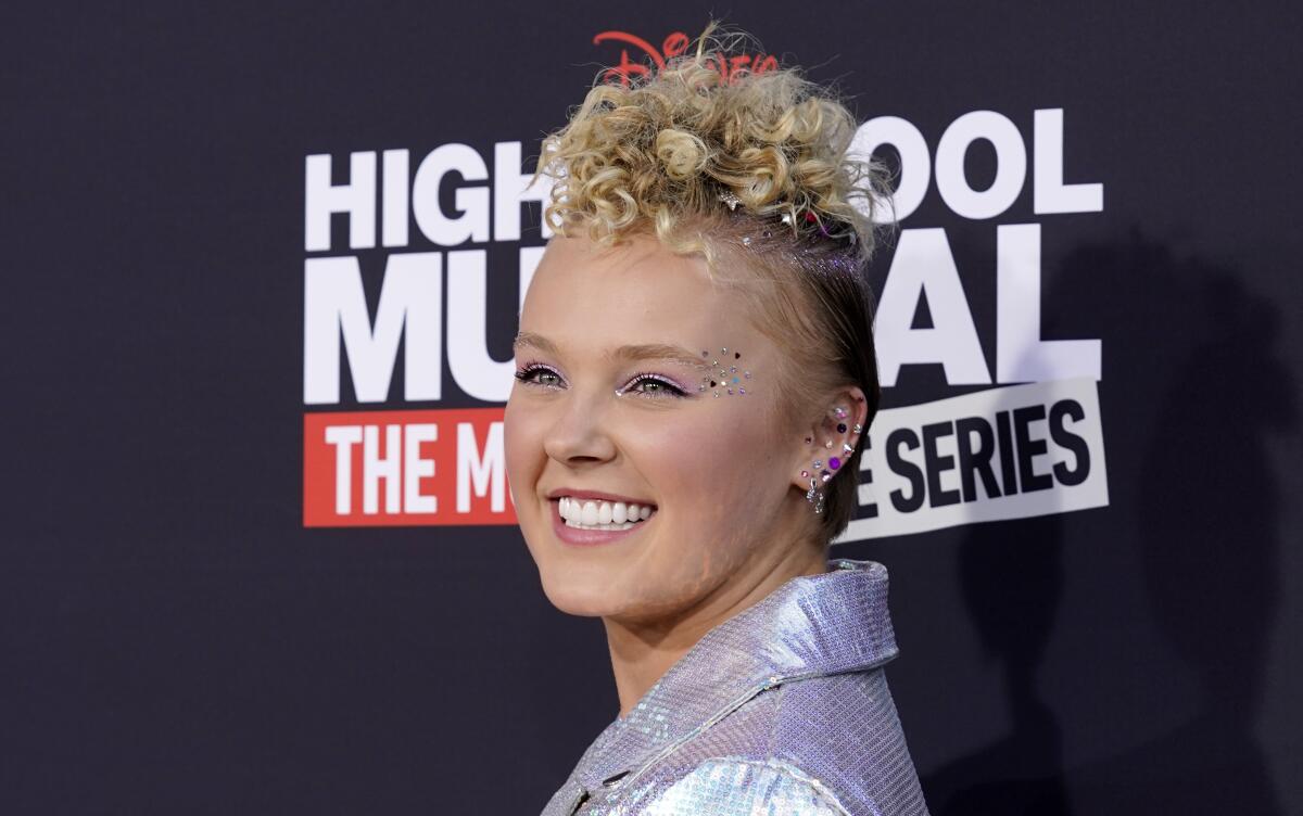 JoJo Siwa smiles while wearing a reflective blazer and sequin makeup at a red carpet event