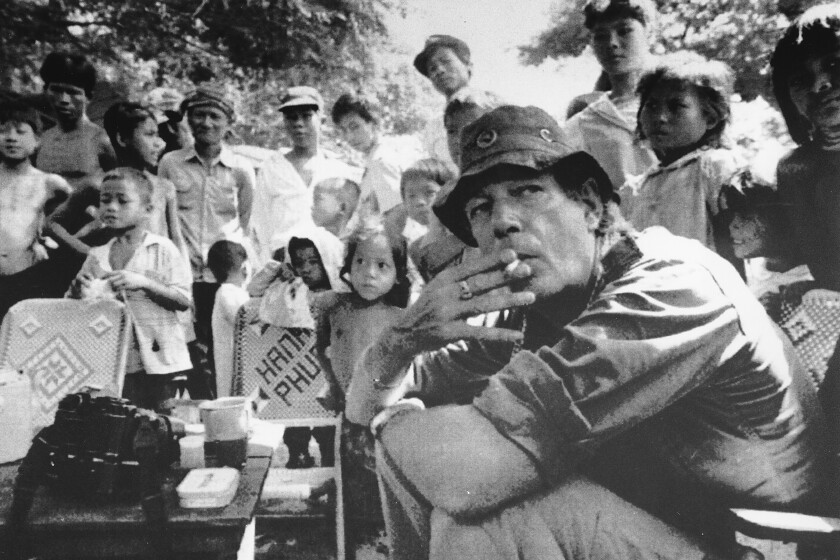 A black and white photo shows a man seated on the ground and smoking a cigarette while surrounded by children.