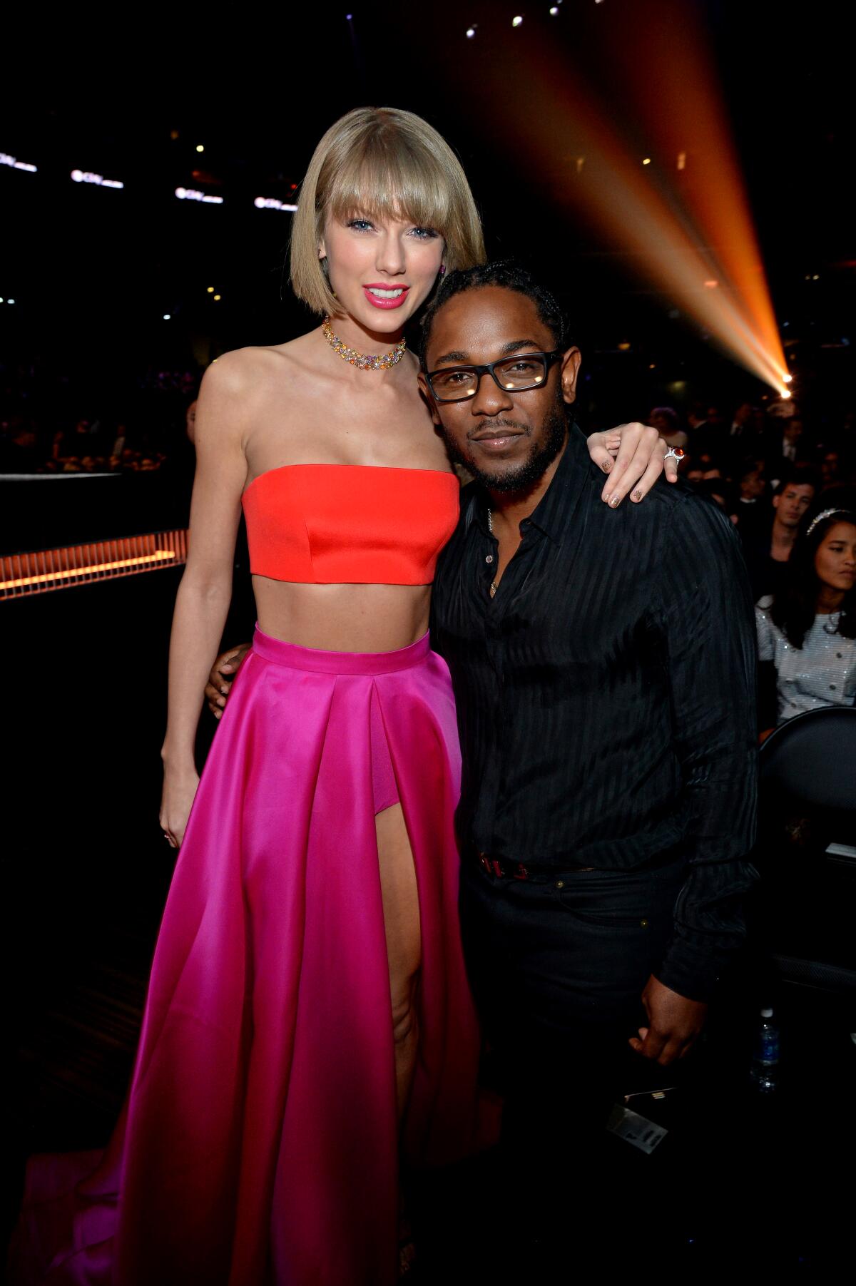 Taylor Swift in a neon orange top and hot pink skirt standing next to Kendrick Lamar in glasses and a black outfit