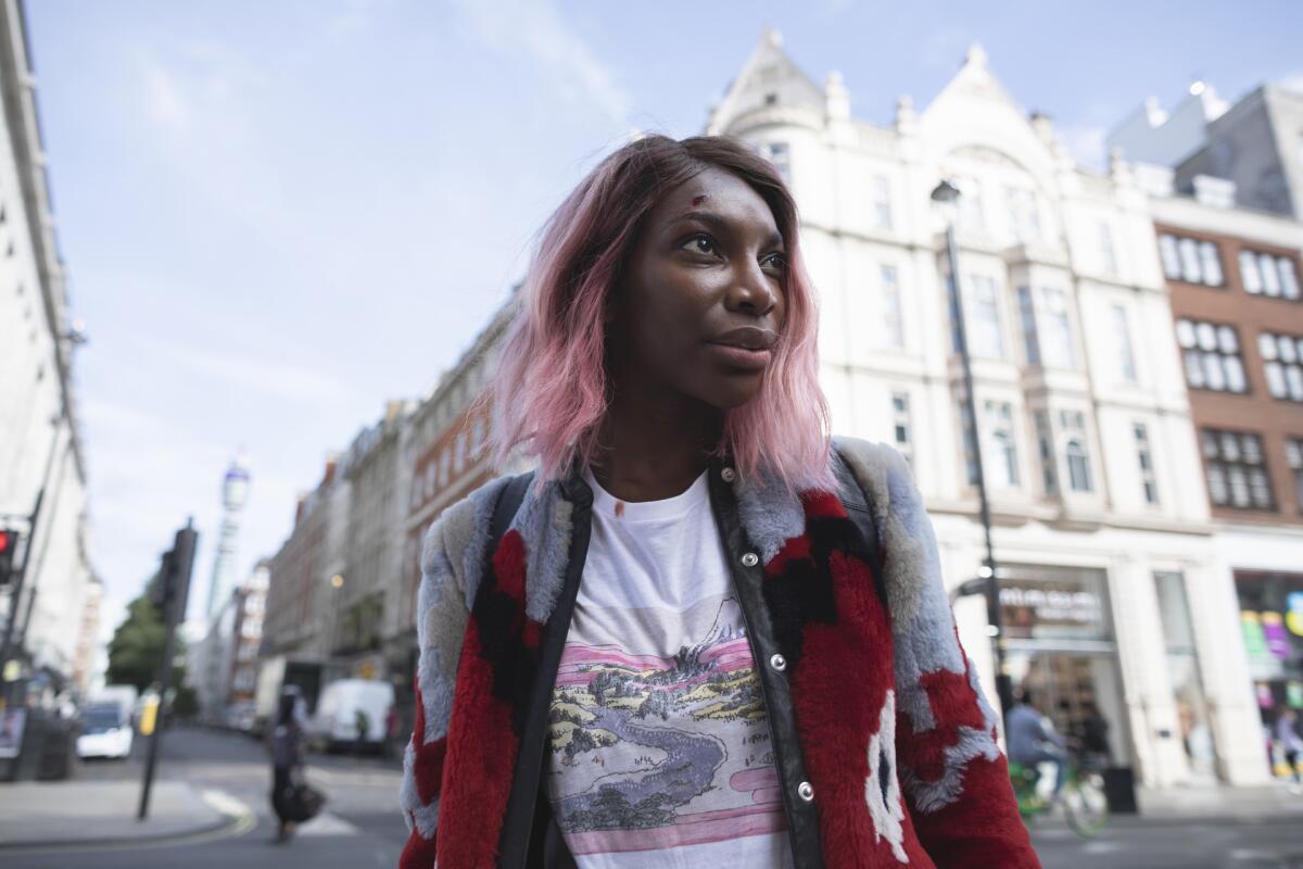 A woman with pink hair and cardigan stands on a city street.