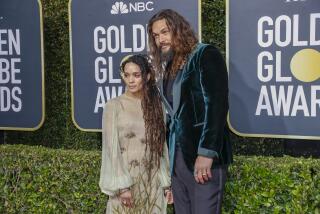 Lisa Bonet wearing a sheer green dress with floral patterns poses next to Jason Momoa wearing a suede green suit jacket