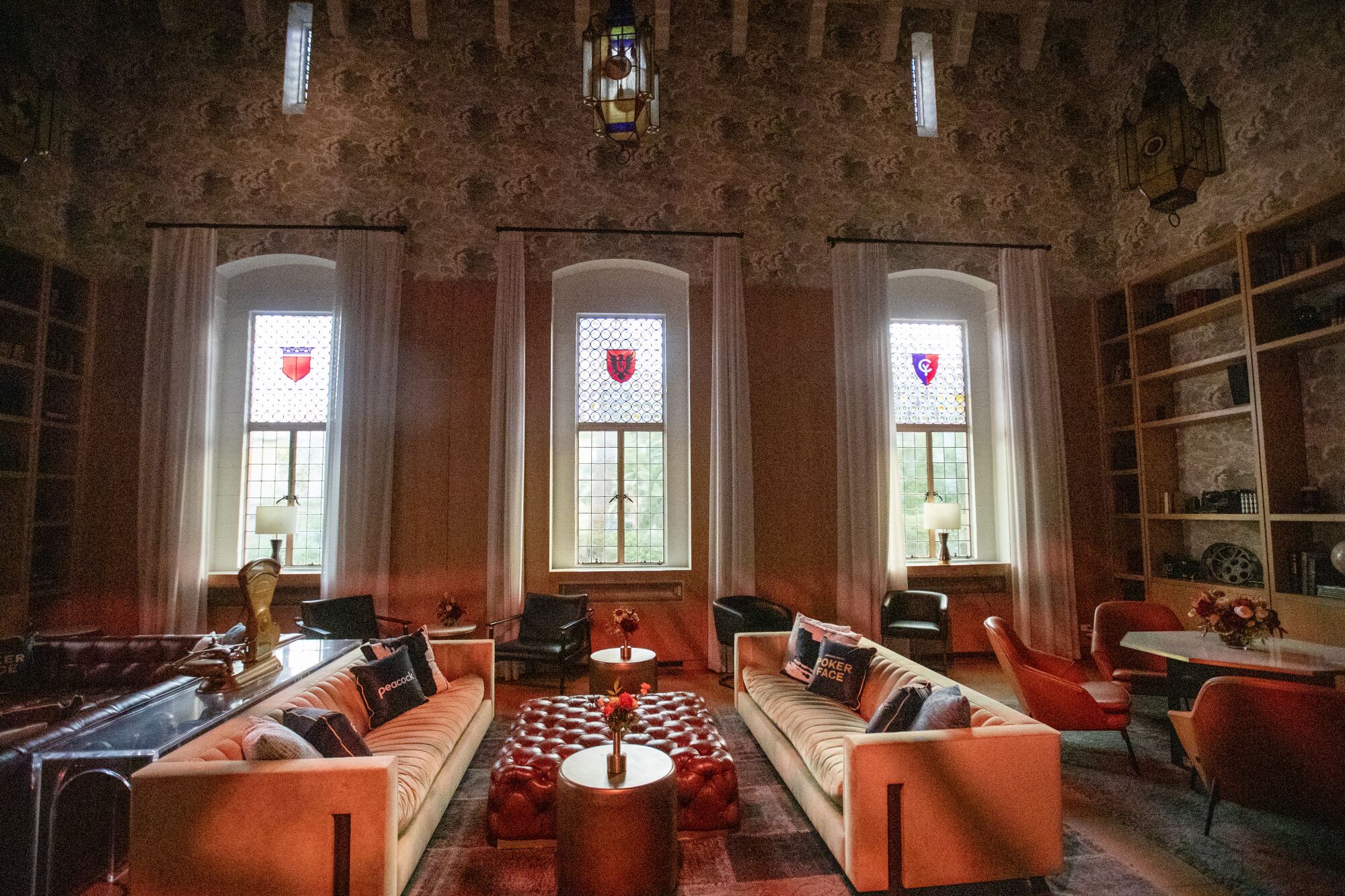 A room with long couches, a variety of chairs and three vertical windows with crests on them