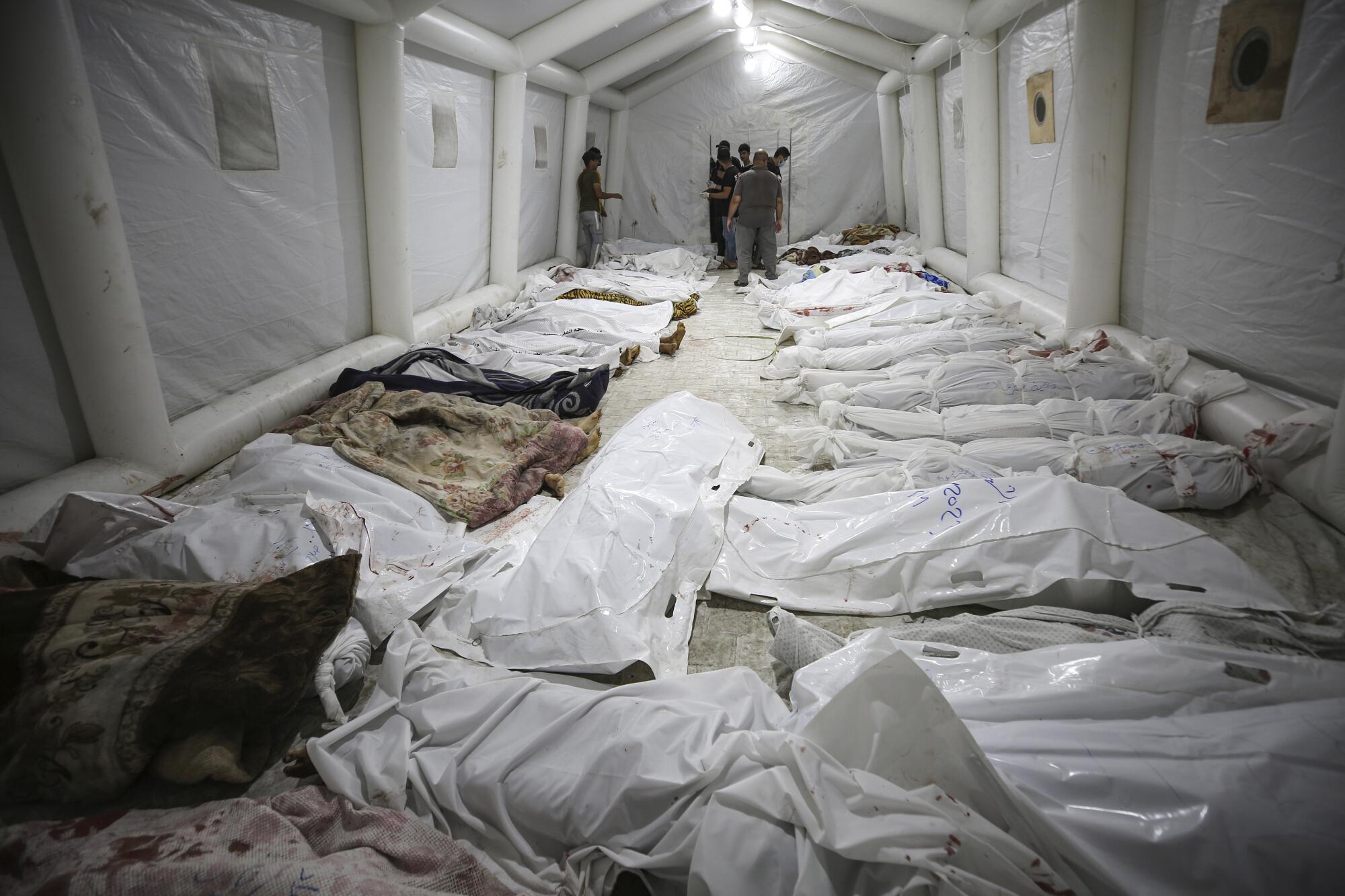 Bodies wrapped in white cloth lie on the floor of a room with white walls 