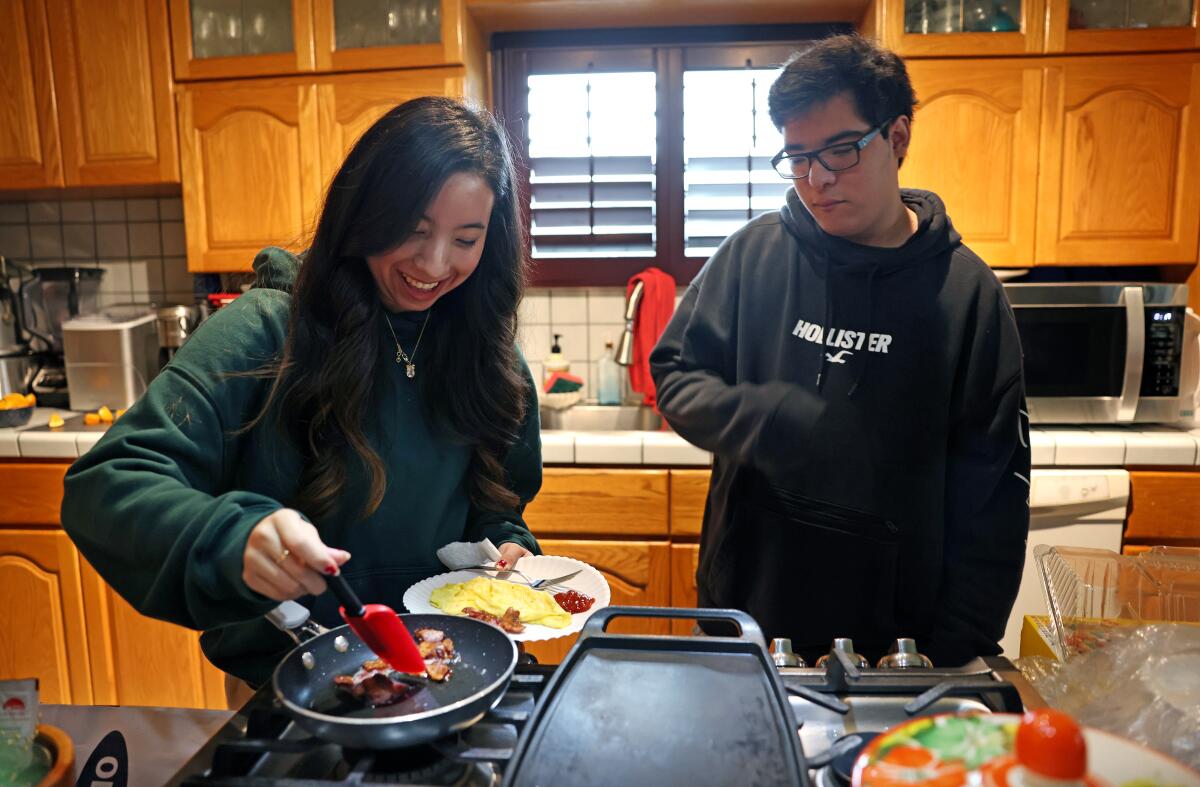 A young woman makes bacon and eggs on a stove as a young man looks on