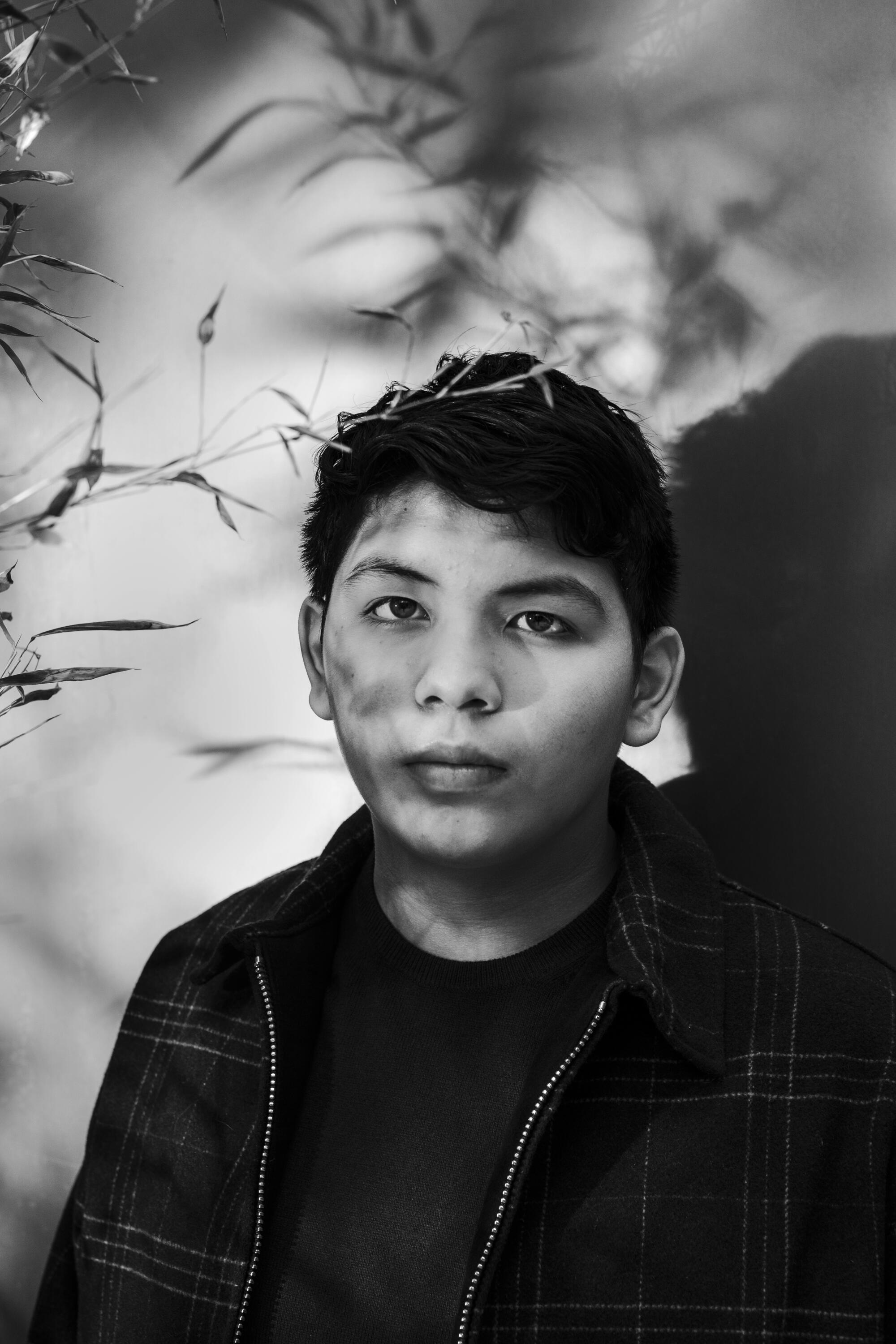 A young man in black poses for a portrait near a cluster of leaves and branches.