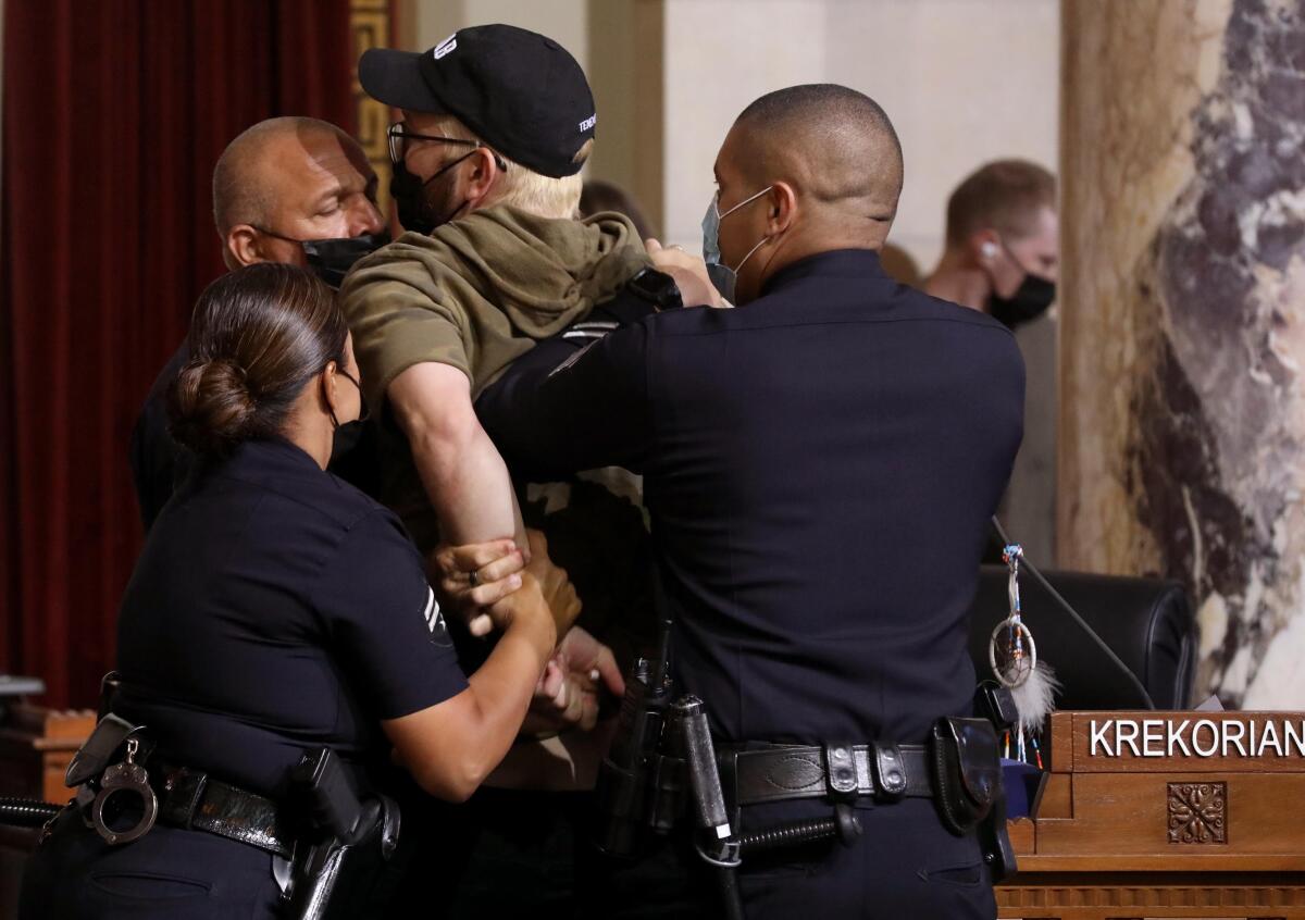 Three police officers restrain a man in a ball cap and face mask.