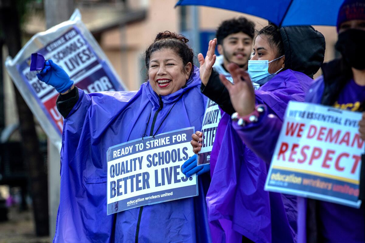 A small group of people, all wearing purple rain coats, hold signs and wave.